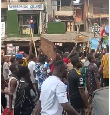 Traders gathered outside barricaded gates of Onitsha Main Market holding handwritten placards, voices raised in protest against the one-week shutdown order; security personnel visible in background under morning sun.
