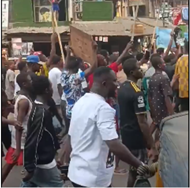 Traders gathered outside barricaded gates of Onitsha Main Market holding handwritten placards, voices raised in protest against the one-week shutdown order; security personnel visible in background under morning sun.